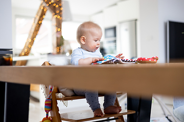 Image showing Happy infant sitting at dining table and playing with his toy in traditional scandinavian designer wooden high chair in modern bright atic home. Cute baby playing with toys