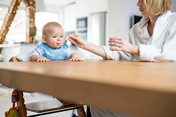 Image showing Mother wearing cosy bathrope spoon feeding her baby boy child in baby high chair with fruit puree at dinning table at home. Baby solid food introduction concept