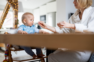 Image showing Mother wearing cosy bathrope spoon feeding her baby boy child in baby high chair with fruit puree at dinning table at home. Baby solid food introduction concept