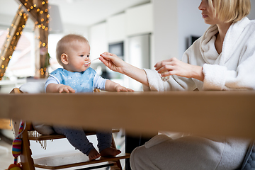 Image showing Mother wearing cosy bathrope spoon feeding her baby boy child in baby high chair with fruit puree at dinning table at home. Baby solid food introduction concept