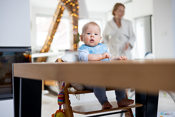 Image showing Happy infant sitting at dining table and playing with his toy in traditional scandinavian designer wooden high chair in modern bright atic home superwised by his mother.