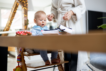 Image showing Happy infant sitting at dining table and playing with his toy in traditional scandinavian designer wooden high chair in modern bright atic home superwised by his mother