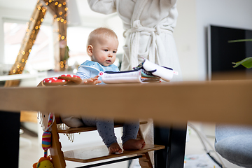 Image showing Happy infant sitting at dining table and playing with his toy in traditional scandinavian designer wooden high chair in modern bright atic home superwised by his mother.