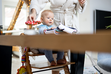 Image showing Happy infant sitting at dining table and playing with his toy in traditional scandinavian designer wooden high chair in modern bright atic home superwised by his mother.