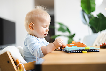 Image showing Happy infant sitting at dining table and playing with his toy in traditional scandinavian designer wooden high chair in modern bright atic home. Cute baby playing with toys