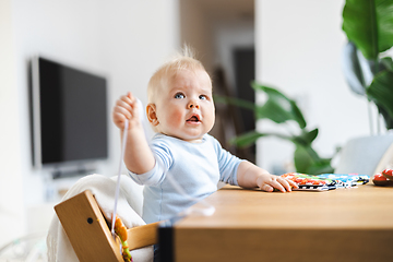 Image showing Happy infant sitting at dining table and playing with his toy in traditional scandinavian designer wooden high chair in modern bright atic home. Cute baby playing with toys