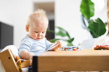 Image showing Happy infant sitting at dining table and playing with his toy in traditional scandinavian designer wooden high chair in modern bright atic home. Cute baby playing with toys