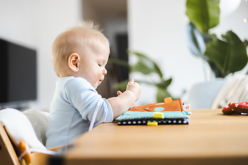 Image showing Happy infant sitting at dining table and playing with his toy in traditional scandinavian designer wooden high chair in modern bright atic home. Cute baby playing with toys