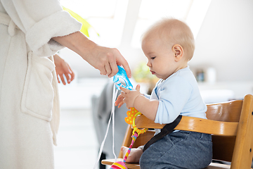 Image showing Happy infant sitting at dining table and playing with his toy in traditional scandinavian designer wooden high chair in modern bright atic home superwised by his mother