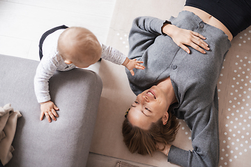 Image showing Happy family moments. Mother lying comfortably on children's mat playing with her baby boy watching and suppervising his first steps. Positive human emotions, feelings, joy.