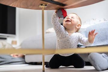 Image showing Cute infant baby boy playing with hanging ball, crawling and standing up by living room table at home. Baby activity and play center for early infant development. Baby playing at home.