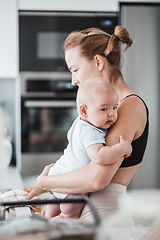 Image showing Woman wiping kitchen sink with a cloth after finishing washing the dishes while holding four months old baby boy in her hands