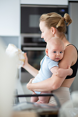 Image showing Woman wiping kitchen sink with a cloth after finishing washing the dishes while holding four months old baby boy in her hands