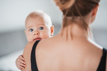 Image showing Portrait of sweet baby resting in mothers arms, looking at camera, touching mama shoulder. New mom holding little kid, embracing child with tenderness, love, care. Motherhood concept