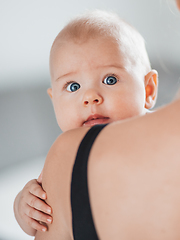 Image showing Portrait of sweet baby resting in mothers arms, looking at camera, touching mama shoulder. New mom holding little kid, embracing child with tenderness, love, care. Motherhood concept