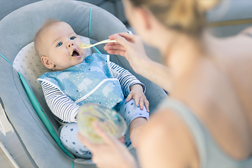 Image showing Mother spoon feeding her baby boy infant child in baby chair with fruit puree. Baby solid food introduction concept.