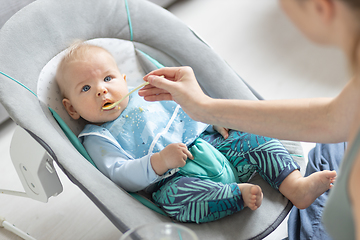 Image showing Mother spoon feeding her baby boy infant child in baby chair with fruit puree. Baby solid food introduction concept.