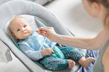 Image showing Mother spoon feeding her baby boy infant child in baby chair with fruit puree. Baby solid food introduction concept.