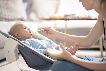 Image showing Mother spoon feeding her baby boy infant child in baby chair with fruit puree. Baby solid food introduction concept.