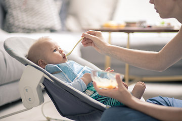 Image showing Mother spoon feeding her baby boy infant child in baby chair with fruit puree. Baby solid food introduction concept.