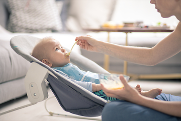 Image showing Mother spoon feeding her baby boy infant child in baby chair with fruit puree. Baby solid food introduction concept.