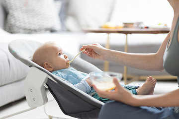 Image showing Mother spoon feeding her baby boy infant child in baby chair with fruit puree. Baby solid food introduction concept.