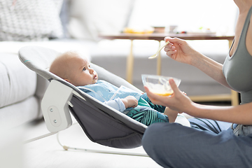 Image showing Mother spoon feeding her baby boy infant child in baby chair with fruit puree. Baby solid food introduction concept.