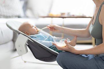 Image showing Mother spoon feeding her baby boy infant child in baby chair with fruit puree. Baby solid food introduction concept.