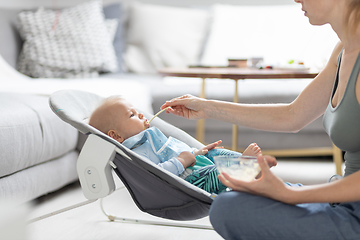 Image showing Mother spoon feeding her baby boy infant child in baby chair with fruit puree. Baby solid food introduction concept.