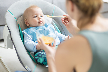 Image showing Mother spoon feeding her baby boy infant child in baby chair with fruit puree. Baby solid food introduction concept.