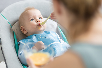 Image showing Mother spoon feeding her baby boy infant child in baby chair with fruit puree. Baby solid food introduction concept.