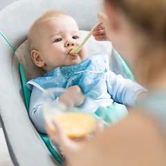 Image showing Mother spoon feeding her baby boy infant child in baby chair with fruit puree. Baby solid food introduction concept.