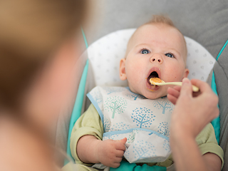 Image showing Mother spoon feeding her baby boy infant child in baby chair with fruit puree. Baby solid food introduction concept.