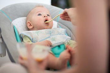 Image showing Mother spoon feeding her baby boy infant child in baby chair with fruit puree. Baby solid food introduction concept.