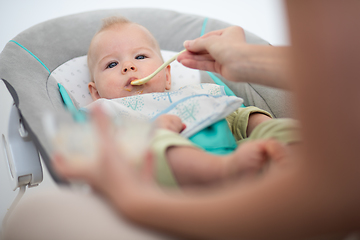 Image showing Mother spoon feeding her baby boy infant child in baby chair with fruit puree. Baby solid food introduction concept.