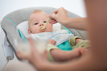 Image showing Mother spoon feeding her baby boy infant child in baby chair with fruit puree. Baby solid food introduction concept.