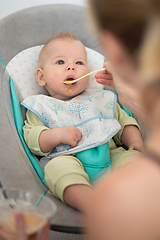 Image showing Mother spoon feeding her baby boy infant child in baby chair with fruit puree. Baby solid food introduction concept.