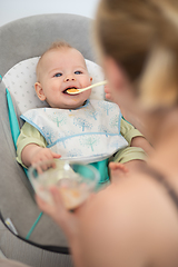 Image showing Mother spoon feeding her baby boy infant child in baby chair with fruit puree. Baby solid food introduction concept.