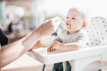 Image showing Mother spoon feeding her baby boy child in baby chair with fruit puree on a porch on summer vacations. Baby solid food introduction concept.