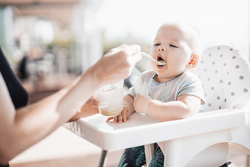 Image showing Mother spoon feeding her baby boy child in baby chair with fruit puree on a porch on summer vacations. Baby solid food introduction concept.