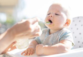 Image showing Mother spoon feeding her baby boy child in baby chair with fruit puree on a porch on summer vacations. Baby solid food introduction concept.