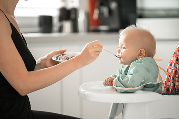 Image showing Mother spoon feeding her baby boy child in baby chair with fruit puree in kitchen at home. Baby solid food introduction concept.