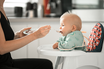 Image showing Mother spoon feeding her baby boy child in baby chair with fruit puree in kitchen at home. Baby solid food introduction concept.