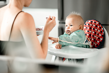 Image showing Mother spoon feeding her baby boy child in baby chair with fruit puree in kitchen at home. Baby solid food introduction concept.