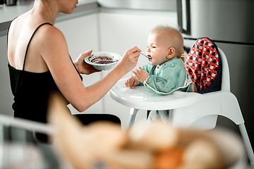 Image showing Mother spoon feeding her baby boy child in baby chair with fruit puree in kitchen at home. Baby solid food introduction concept.