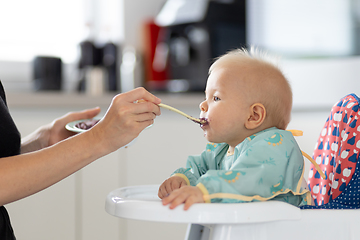 Image showing Mother spoon feeding her baby boy child in baby chair with fruit puree in kitchen at home. Baby solid food introduction concept.