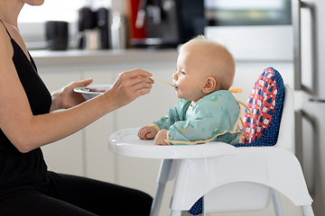 Image showing Mother spoon feeding her baby boy child in baby chair with fruit puree in kitchen at home. Baby solid food introduction concept.