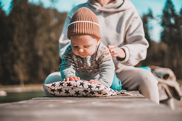 Image showing Happy family. Young mother playing with her baby boy infant oudoors on sunny autumn day. Portrait of mom and little son on wooden platform by lake. Positive human emotions, feelings, joy.