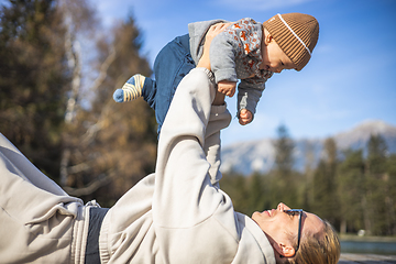 Image showing Happy family. Young mother playing with her baby boy infant oudoors on sunny autumn day. Portrait of mom and little son on wooden platform by lake. Positive human emotions, feelings, joy.