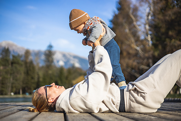 Image showing Happy family. Young mother playing with her baby boy infant oudoors on sunny autumn day. Portrait of mom and little son on wooden platform by lake. Positive human emotions, feelings, joy.